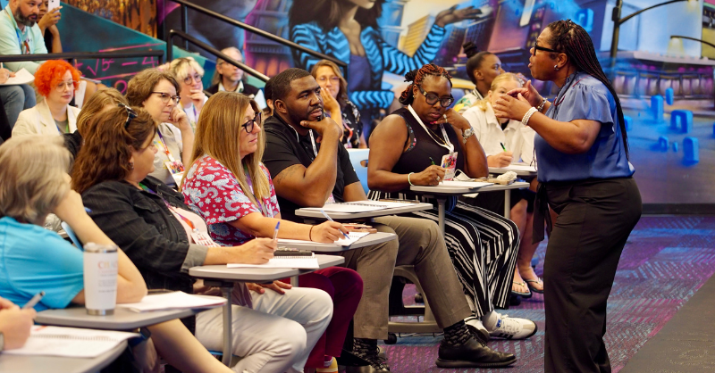 male teacher sitting in desk learning from presenter